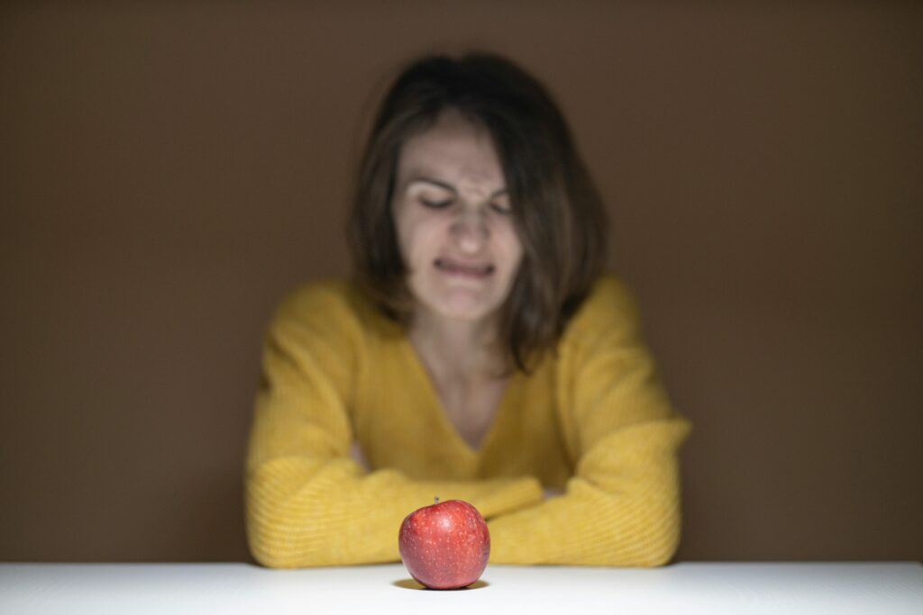 pexels photo 3214347 3214347 A woman in a yellow sweater shows disgust while looking at a fresh red apple on a table.